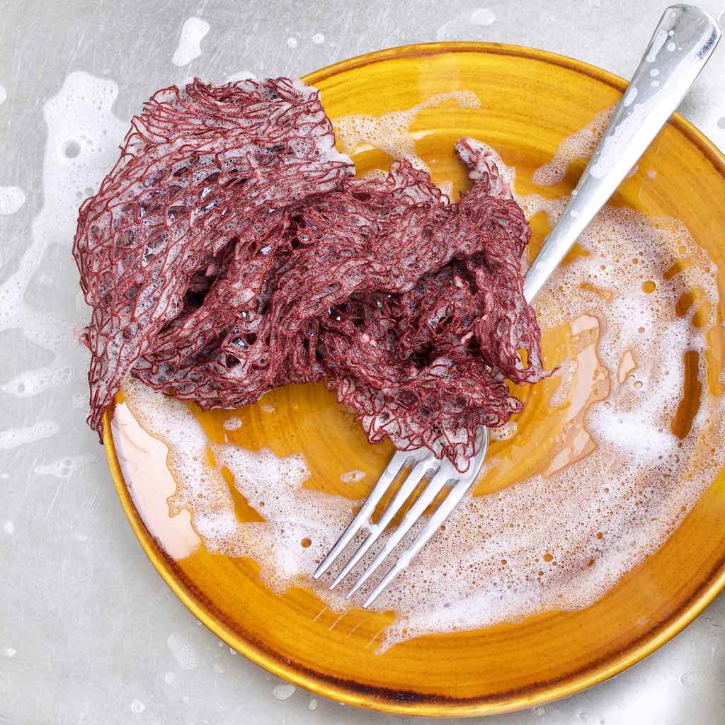 Wet and foamy Fischnetz With Benefits as a dish sponge alternative lying on a brown plate in a kitchen sink.